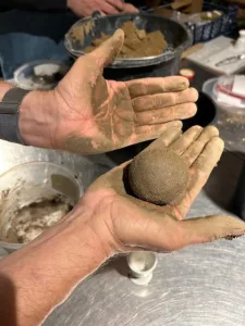 Zwei lehmverschmierte Hände halten eine frisch geformte Dorodango-Kugel aus Erde