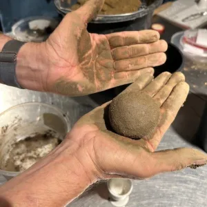Zwei lehmverschmierte H&auml;nde halten eine frisch geformte Dorodango-Kugel aus Erde