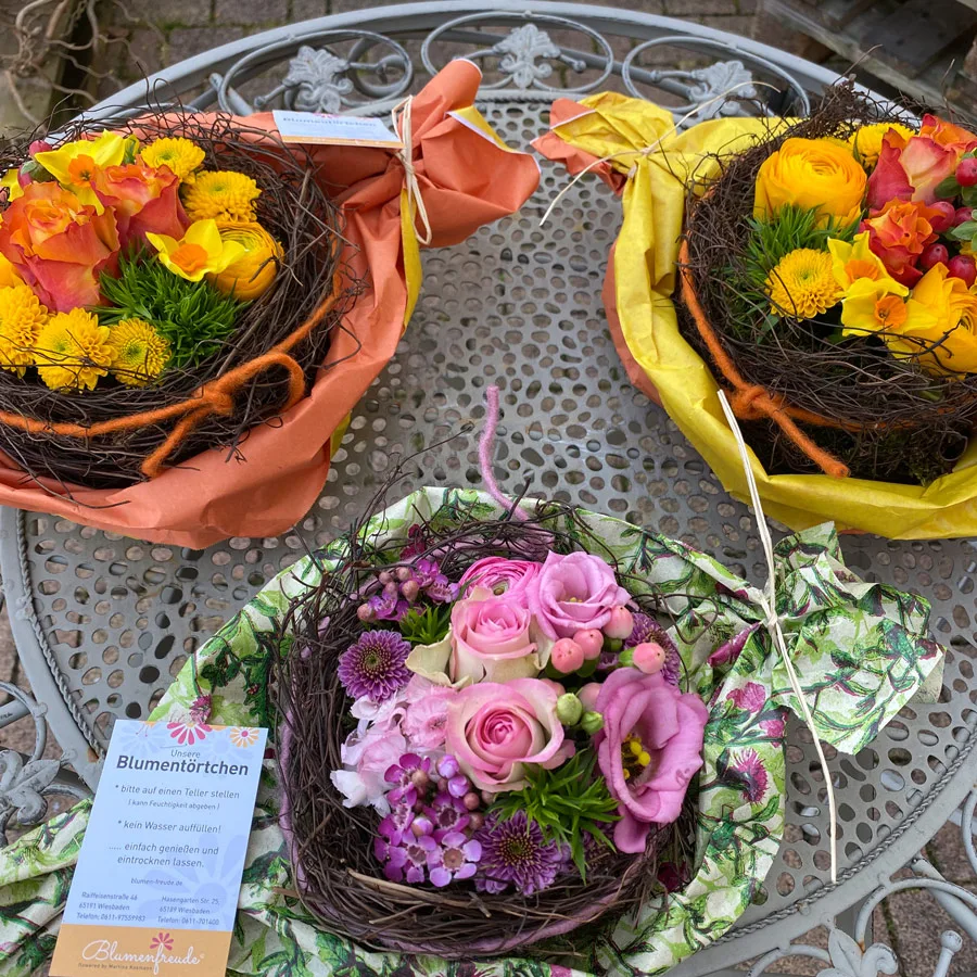 Drei Blumentörtchen in Nestern aus Zweigen mit gelben, orangen und rosa Blüten auf einem Gartentisch