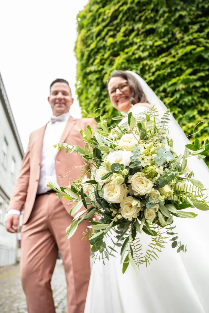 Brautpaar vor einer hohen Hecke, die Braut hält einen weiß-grünen Brautstrauß mit Rosen und Farn