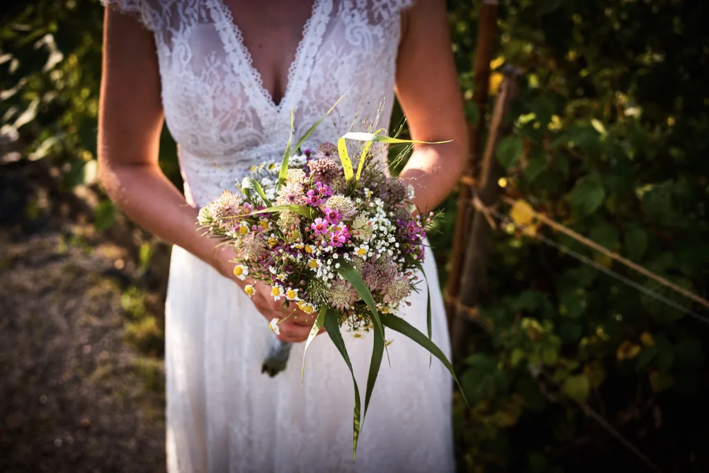 Braut im Spitzenkleid hält einen locker gebundenen Brautstrauß aus rosa und violetten Wiesenblumen
