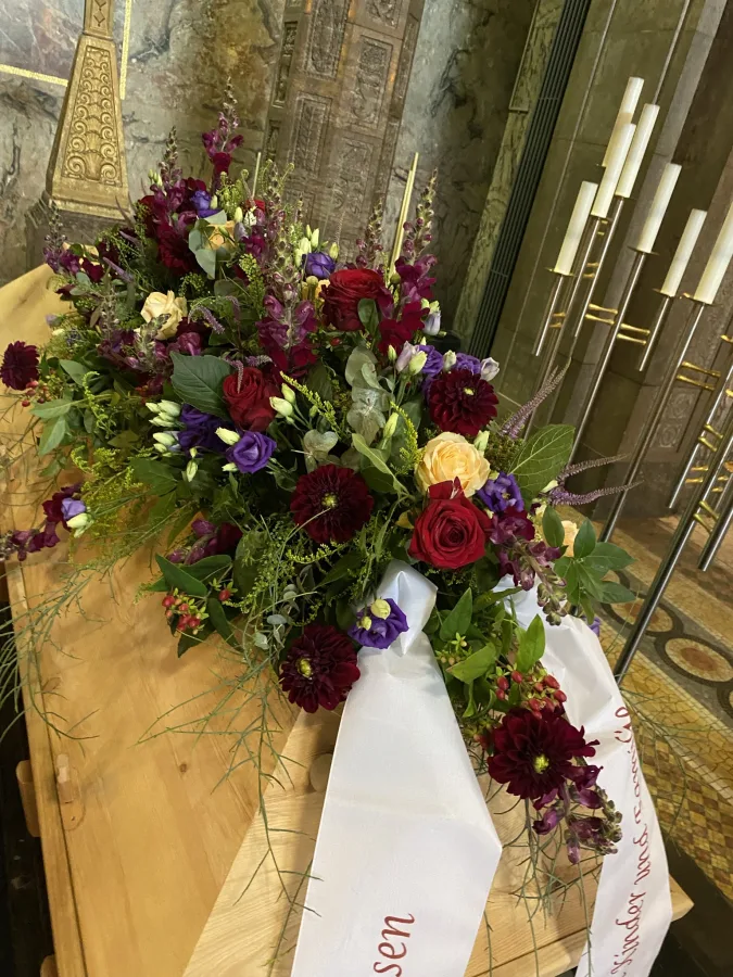 Sargbouquet aus dunkelroten und violetten Blüten mit weißer Schleife auf einem hellen Sarg