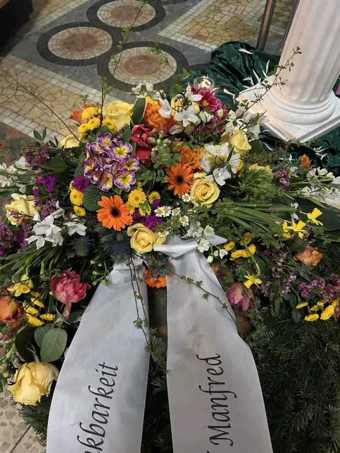 Buntes Trauergesteck mit Gerbera und Rosen und beschrifteten Trauerschleifen