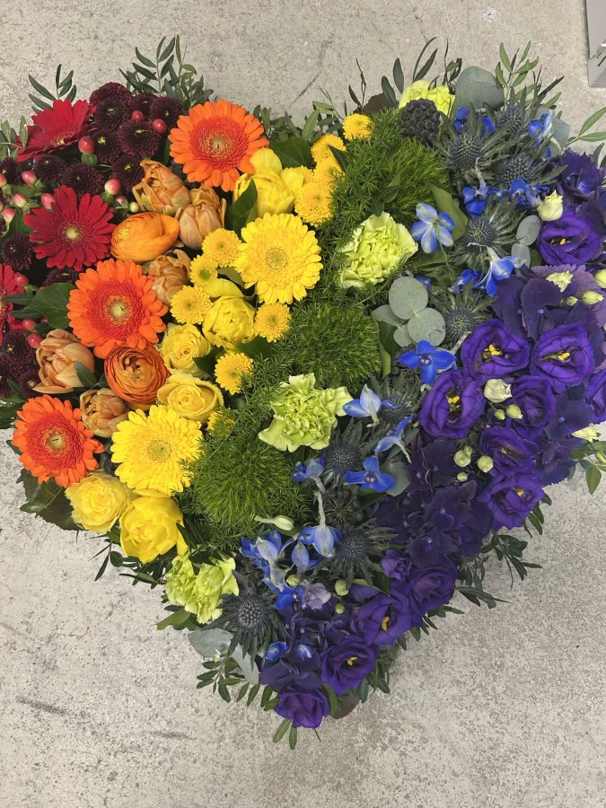 Herzförmiges Trauergesteck mit orangen, gelben, blauen und violetten Blüten