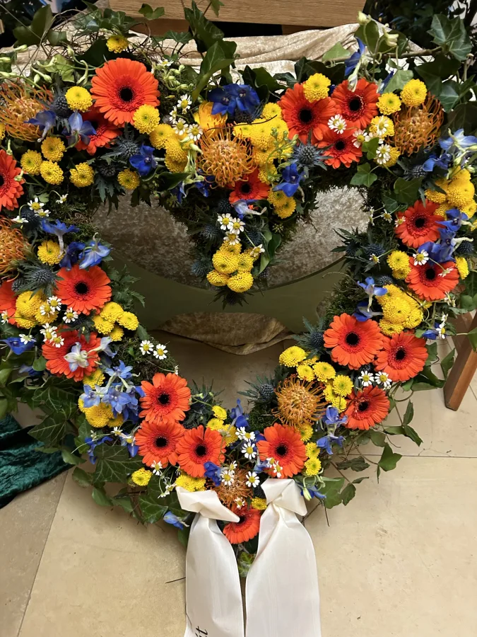 Herzförmiges Trauergesteck mit gelben und orangen Gerbera und blauen Blüten