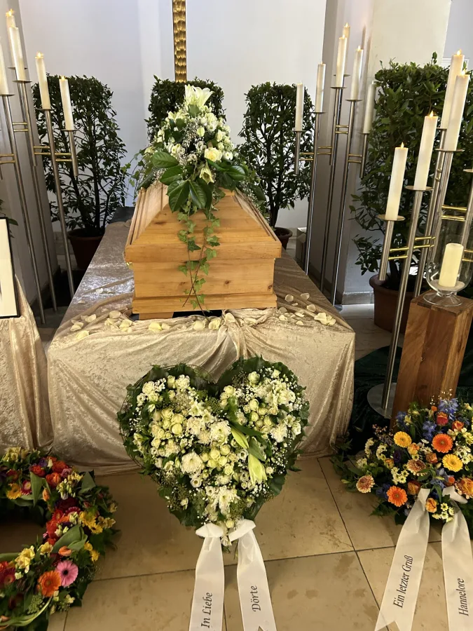 Heller Sarg mit weißem Bouquet und herzförmigem Trauergesteck davor in einer Trauerhalle