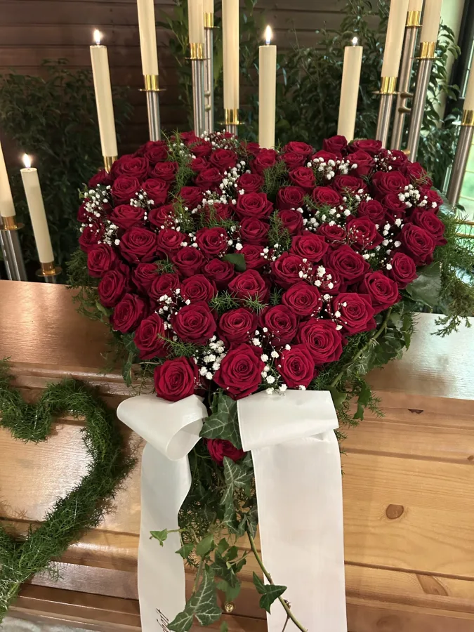 Herzförmiges Trauergesteck aus roten Rosen mit weißen Schleifen vor hohen Kerzenleuchtern