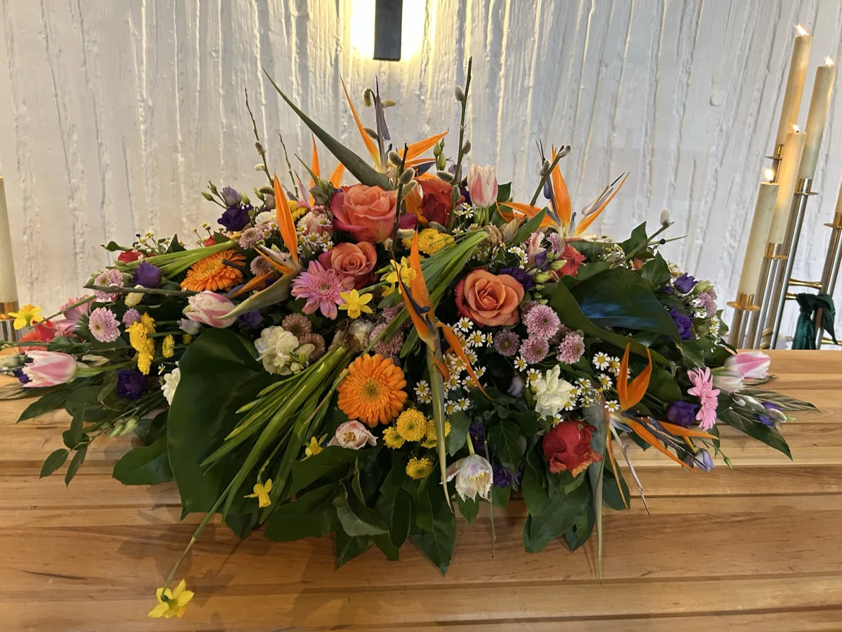 Farbenfrohes Sargbouquet mit Strelitzien, orangen Rosen und Gerbera auf einem Holzsarg