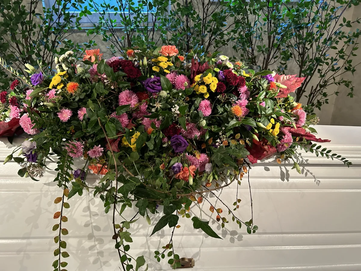 Sargbouquet mit pinken und bordeauxfarbenen Blüten und hängendem Efeu auf einem weißen Sarg