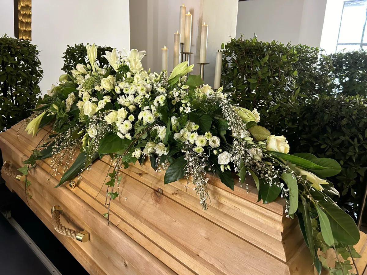 Sargbouquet mit weißen Lilien, Rosen und Eukalyptus auf einem Holzsarg