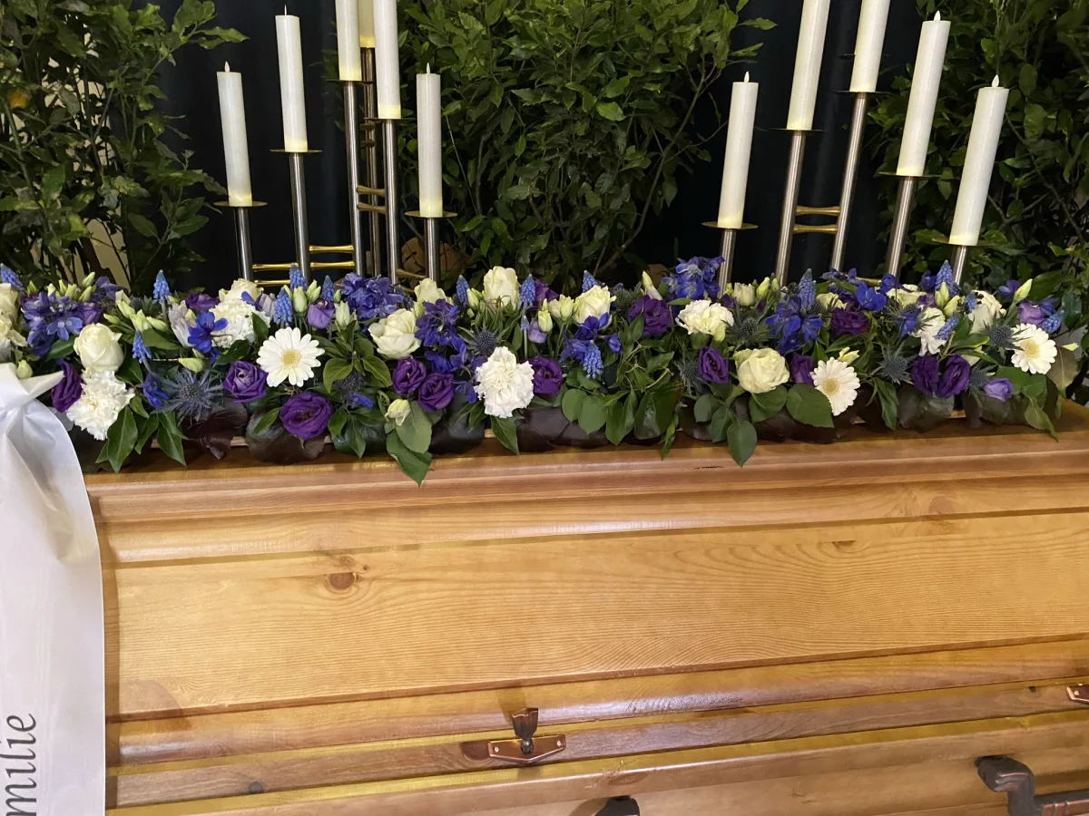 Langgestrecktes Sargbouquet mit blauen und weißen Blüten auf einem Holzsarg