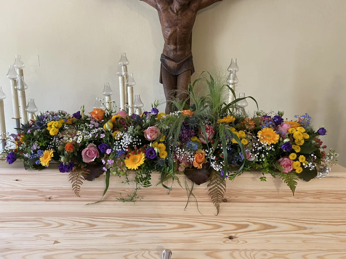 Langgestrecktes Sargbouquet mit bunten Sommerblumen auf einem hellen Holzsarg