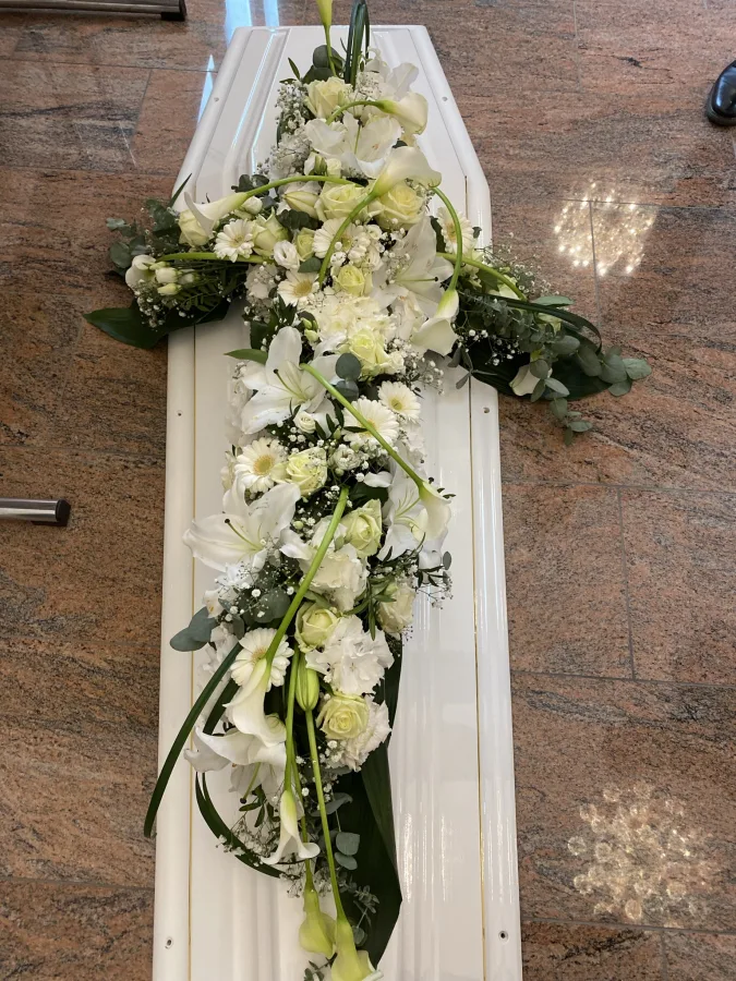 Kreuzförmiges Sargbouquet mit weißen Rosen und Callas auf einem weißen Sarg