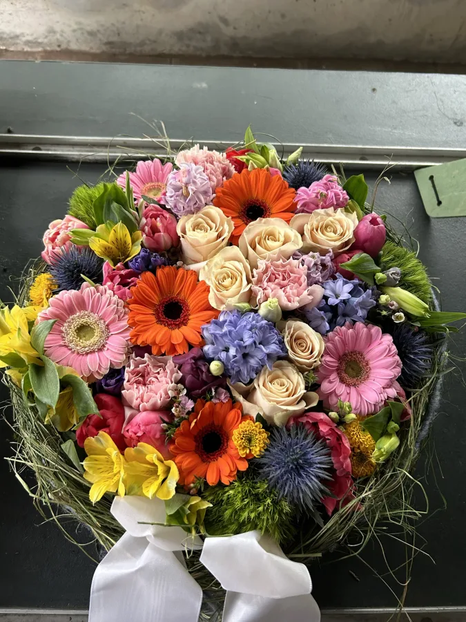 Runder Trauerstrauß mit pinken Gerbera, orangen und violetten Blüten und weißer Schleife