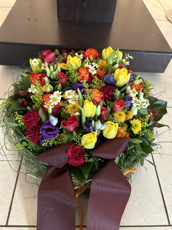 Runder Trauerstrauß mit roten, gelben und violetten Blüten und bordeauxfarbener Schleife