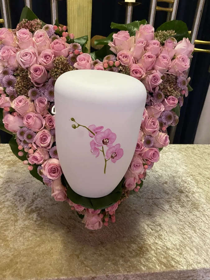 Weiße Urne mit Orchideenmotiv in einem herzförmigen Kranz aus rosa Rosen