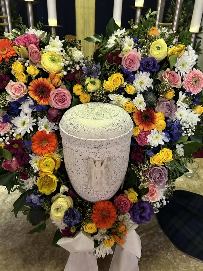 Weiße Urne mit Engelsmotiv, umgeben von bunten Gerbera und Rosen