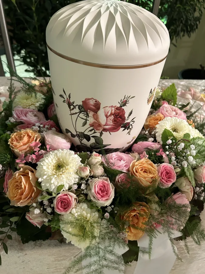 Weiße Urne mit Blumenmotiv, umgeben von rosa Rosen und weißen Chrysanthemen