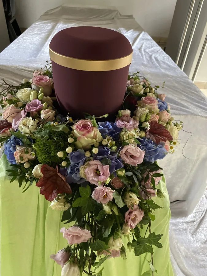 Dunkelrote Urne mit einem Kranz aus rosa Rosen, Efeu und blauen Blüten