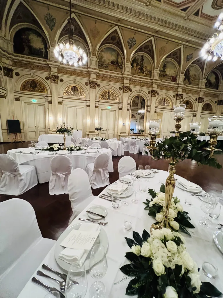 Festlich eingedeckter Saal mit weißen Stuhlhussen und einem goldenen Leuchter mit weißen Rosen